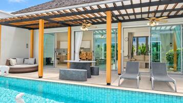 Un área junto a la piscina con zona de estar cubierta, mesa de comedor y sillones reclinables.