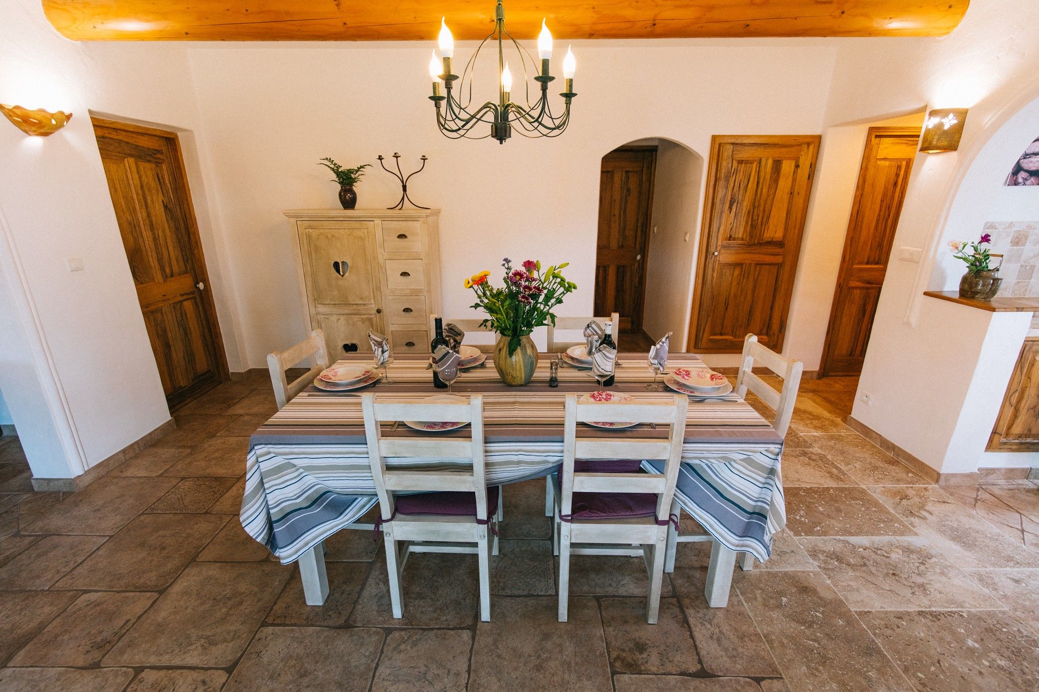 A dining area with a table set for two, wooden chairs, a chandelier, and a vase with flowers.