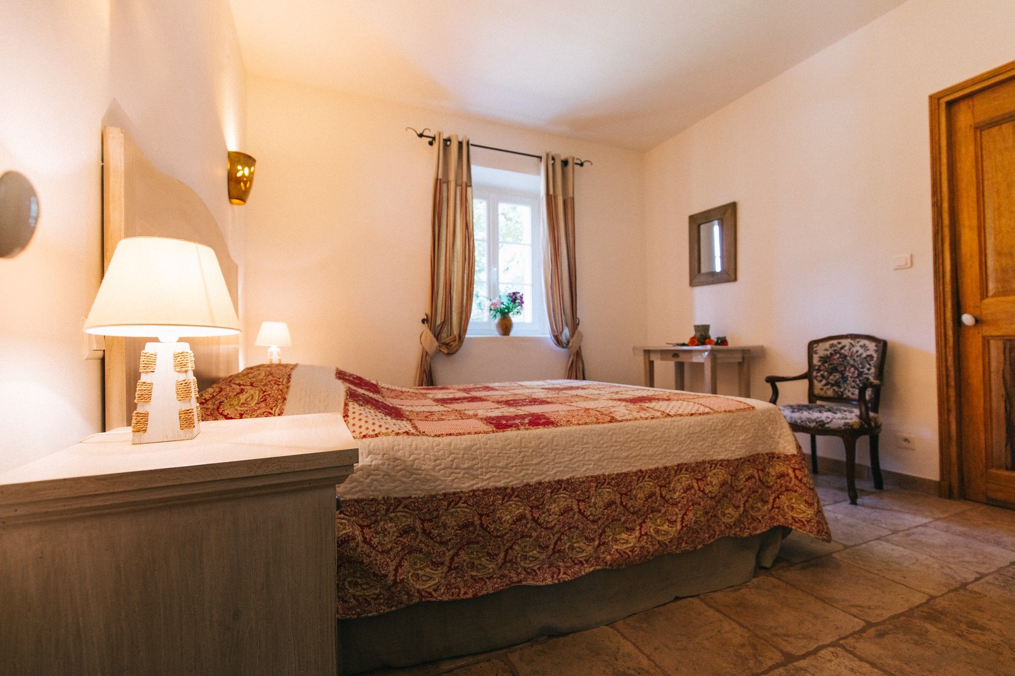 A bedroom with a bed, a bedside table with a lamp, a chair, a window with curtains, and a door.