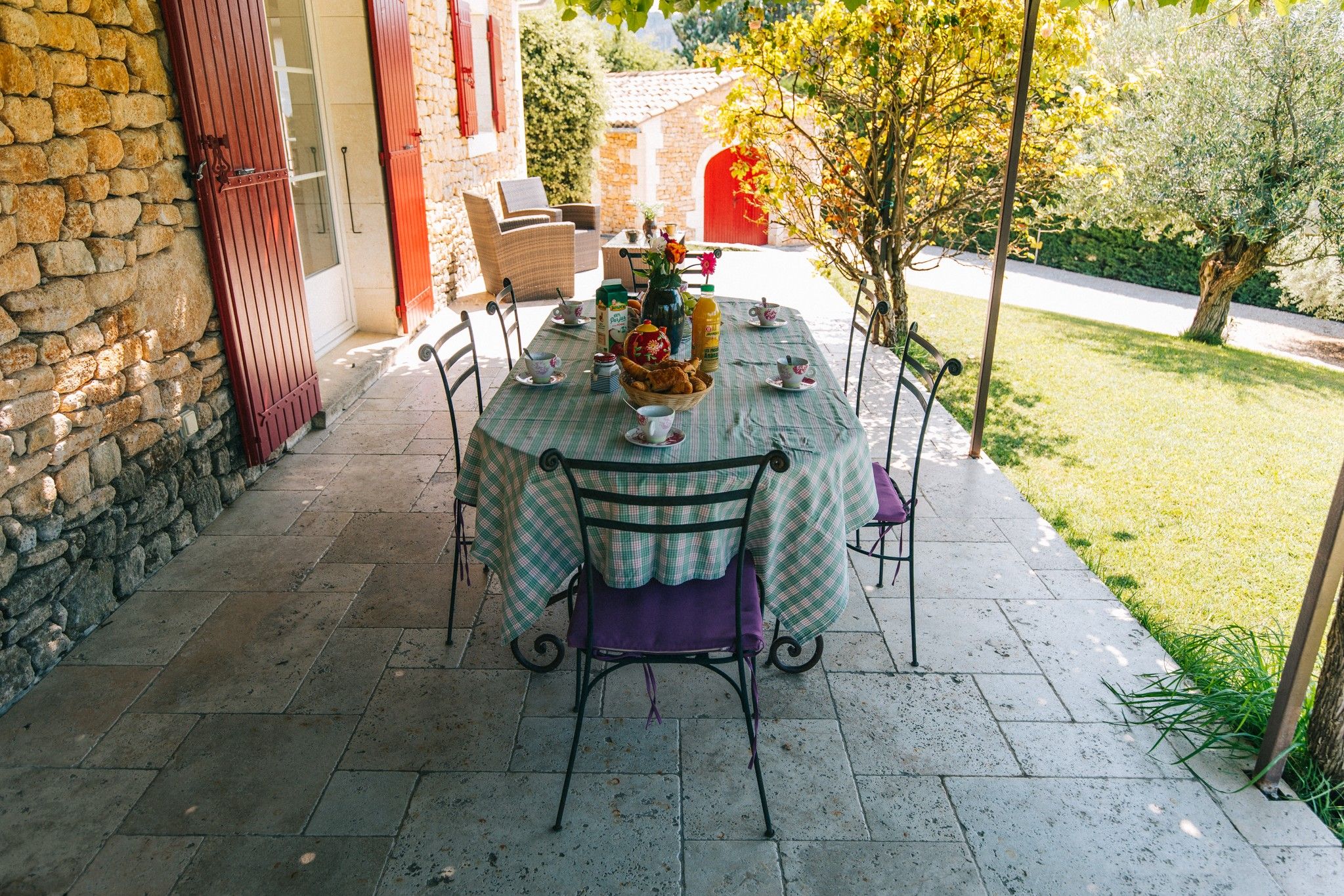 Un espace de restauration en plein air, avec un mur en pierre, une porte rouge et une table dressée avec une nappe à carreaux, des chaises et un panier de pain.