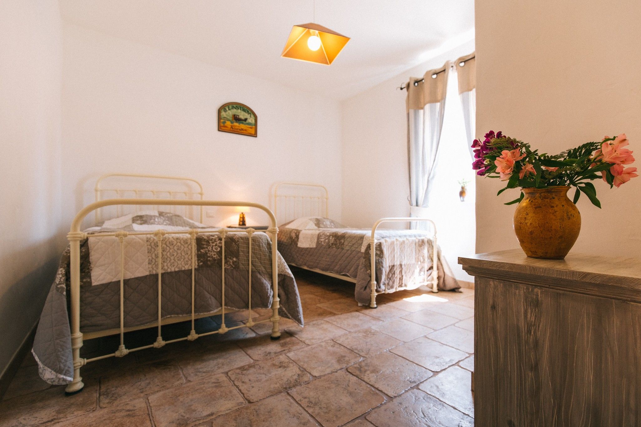 Una habitación con dos camas de metal, una mesita, un jarrón con flores y un cuadro colgado en la pared.