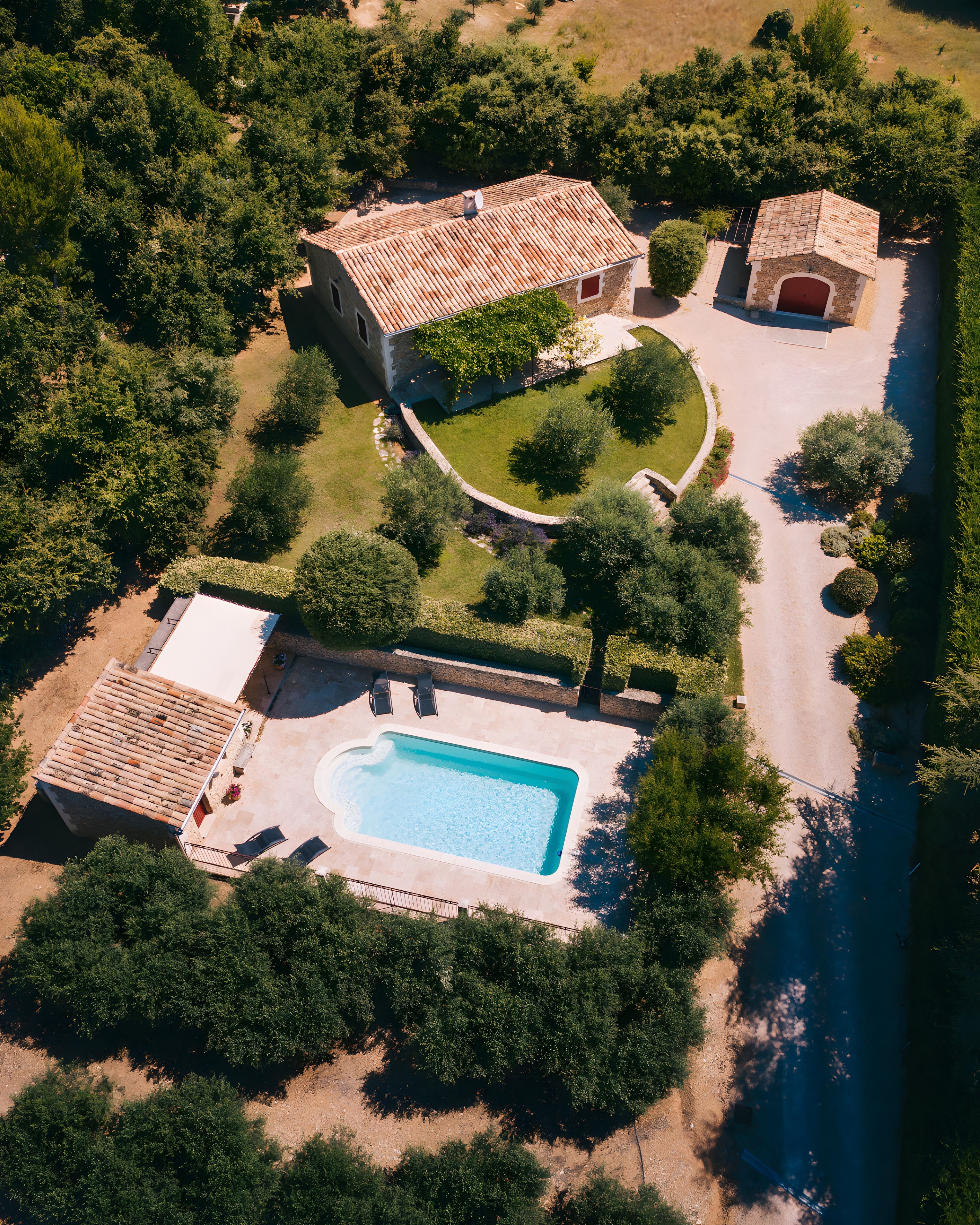 An aerial view of a property with a swimming pool, a stone house with a tiled roof, and surrounding greenery.