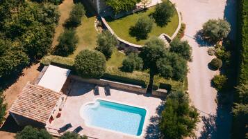 An aerial view of a property with a swimming pool, a stone house with a tiled roof, and surrounding greenery.