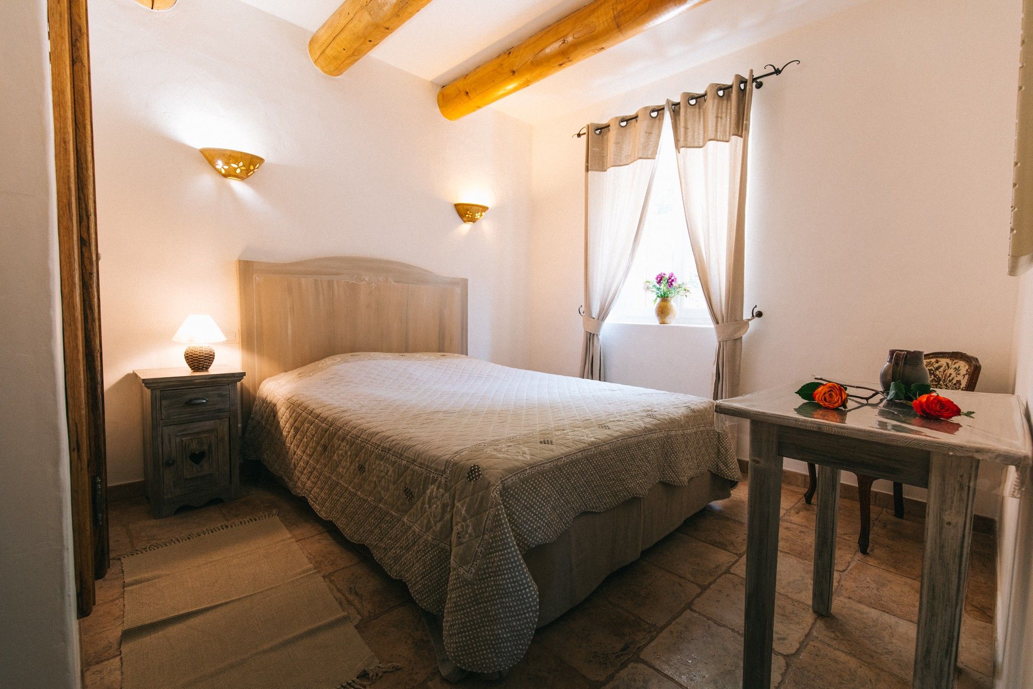 Quarto com cama, mesa de cabeceira, abajur, janela com cortinas e uma pequena mesa com um vaso e flores.