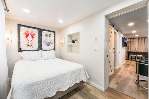 Een slaapkamer met een bed, een wandplank met boeken en een deur naar een gang.