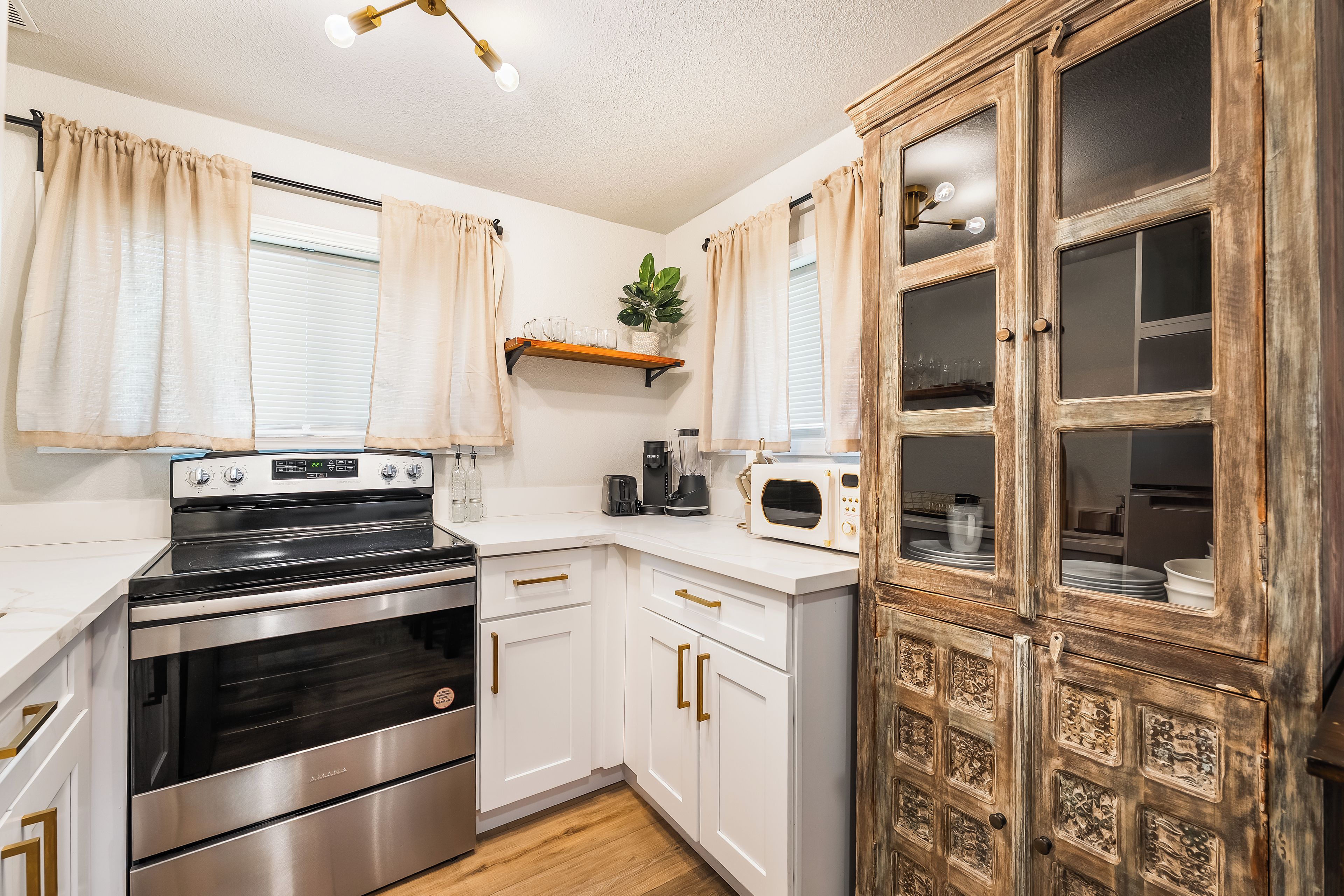 A compact kitchen with white cabinetry, a stainless steel stove, a microwave, and a wooden cabinet with glass doors.