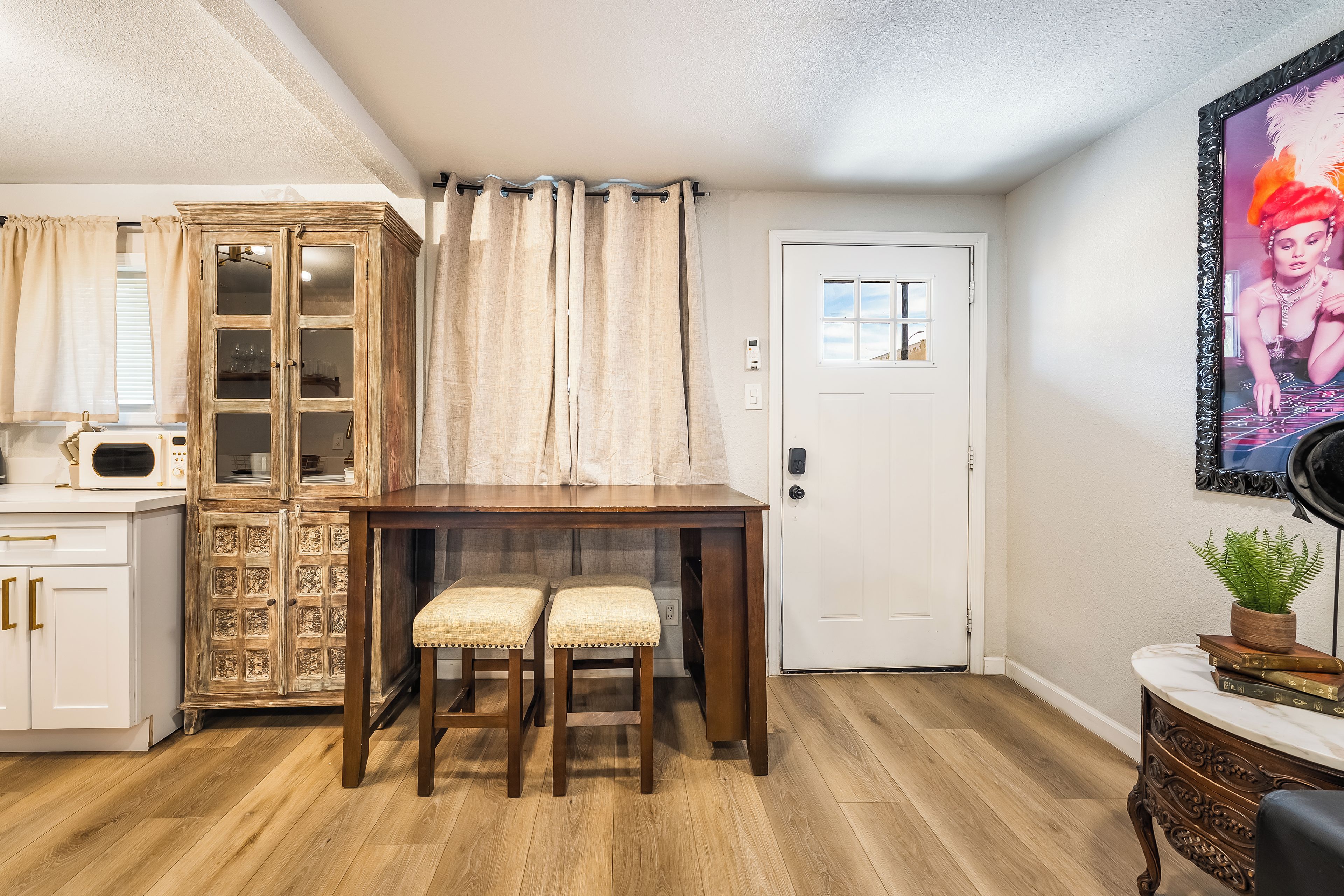 A room with a wooden dining table, two stools, a cabinet with glass doors, a white door, and a framed picture on the wall.