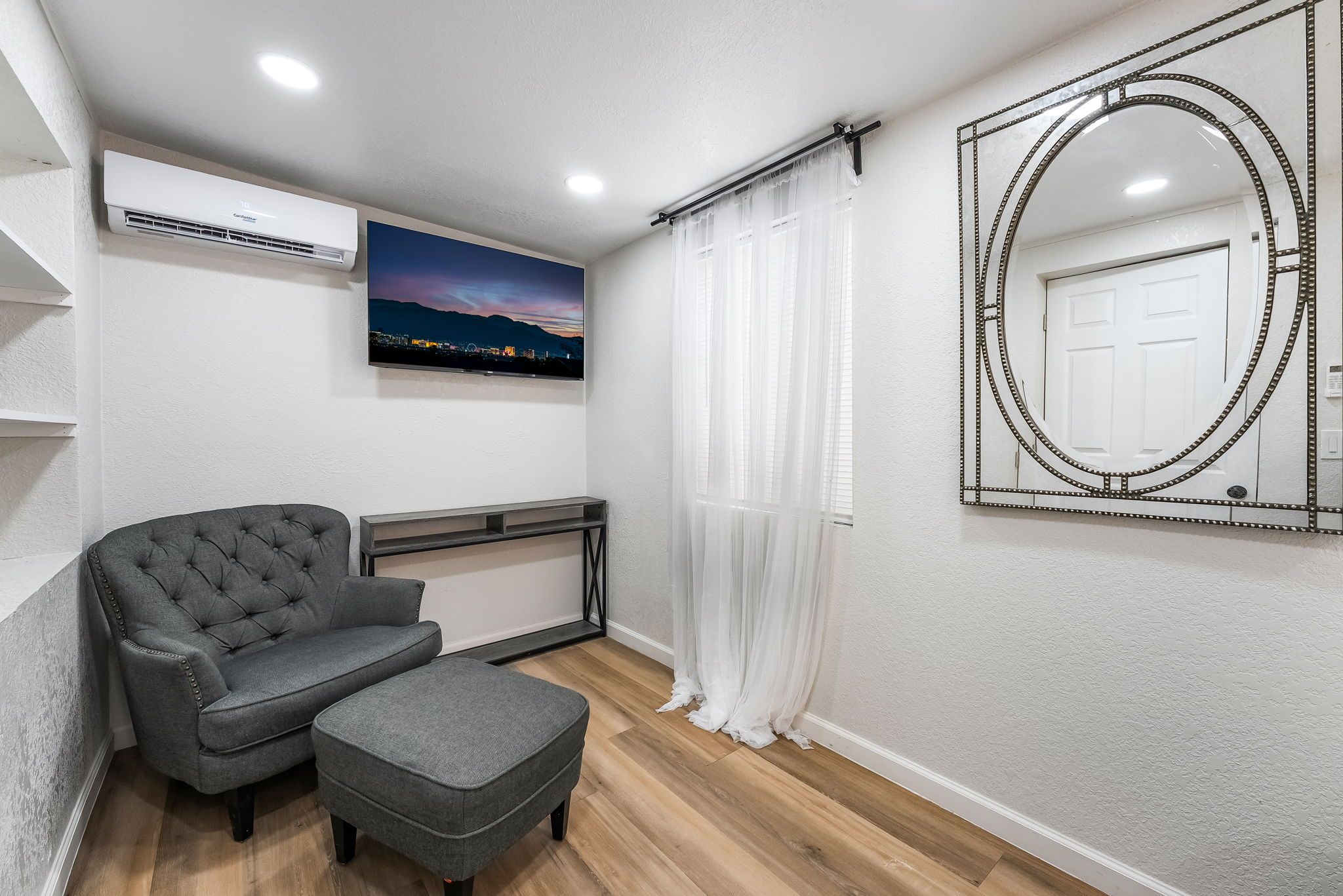 Een moderne kamer met een grijze bank, een klein voetenbankje, een flatscreen televisie en een decoratieve spiegel aan de muur.
