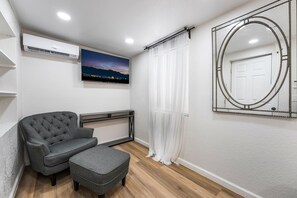 Een moderne kamer met een grijze bank, een klein voetenbankje, een flatscreen televisie en een decoratieve spiegel aan de muur.