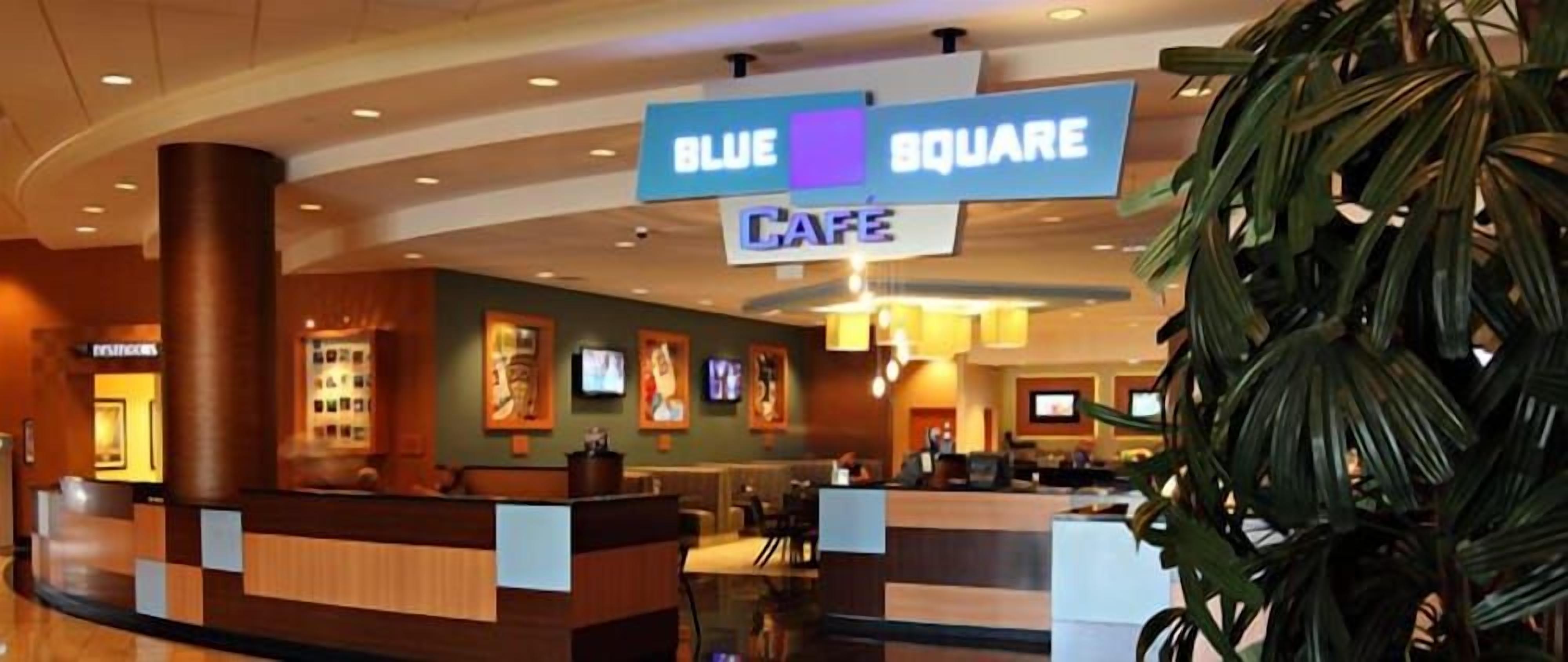 A hotel lobby with a reception desk, a blue sign indicating the Blue Square Cafe, and a large plant in the foreground.