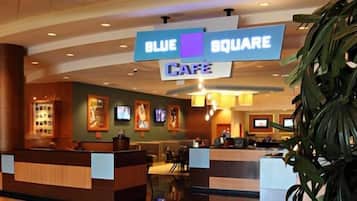 A hotel lobby with a reception desk, a blue sign indicating the Blue Square Cafe, and a large plant in the foreground.