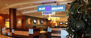 A hotel lobby with a reception desk, a blue sign indicating the Blue Square Cafe, and a large plant in the foreground.