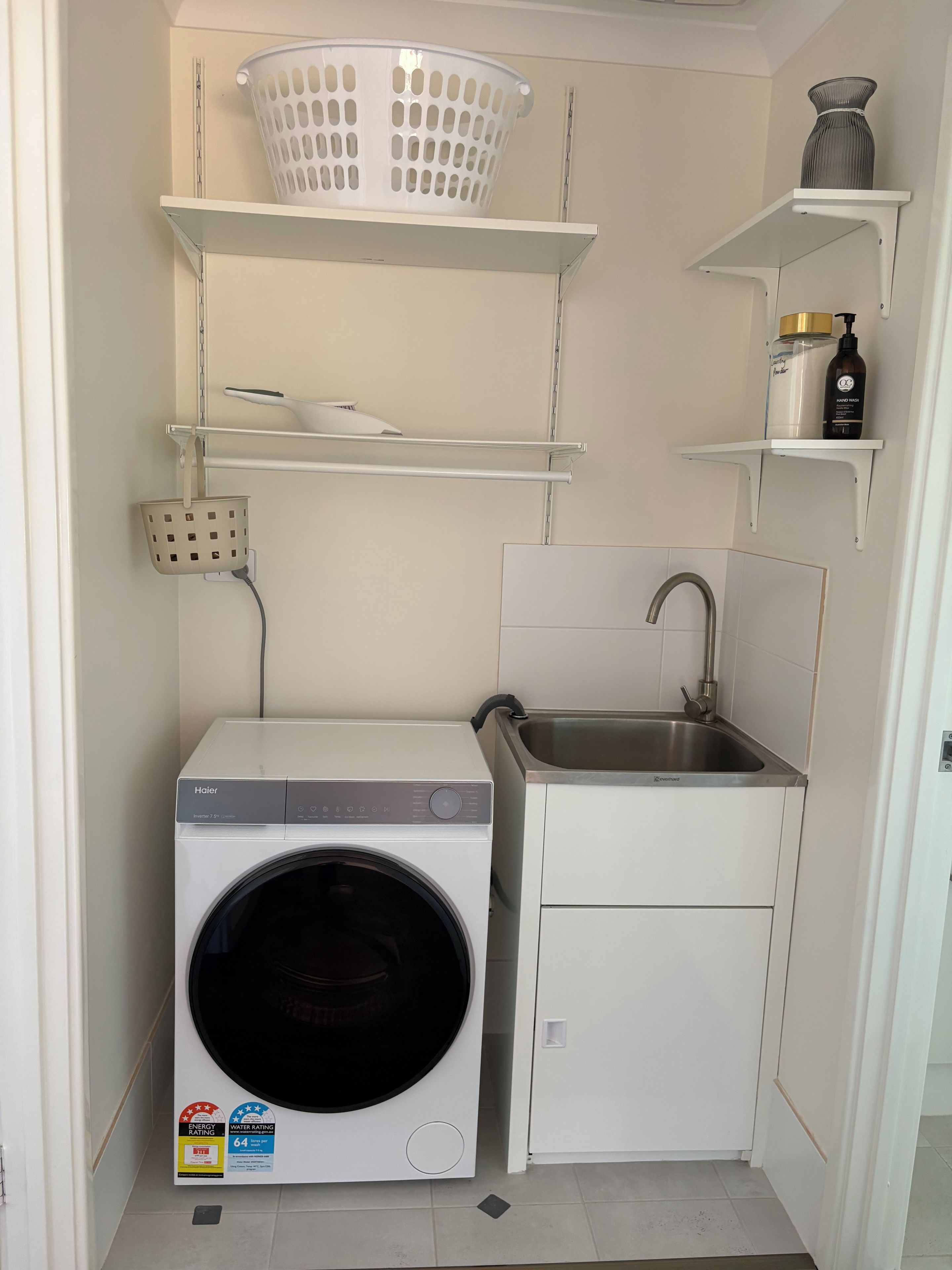 A compact laundry room with a front-loading washer, a sink, and storage shelves.