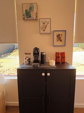 Una kitchenette con cafetera, hervidor y un mueble pequeño. Encima de la encimera hay cuadros enmarcados en la pared.