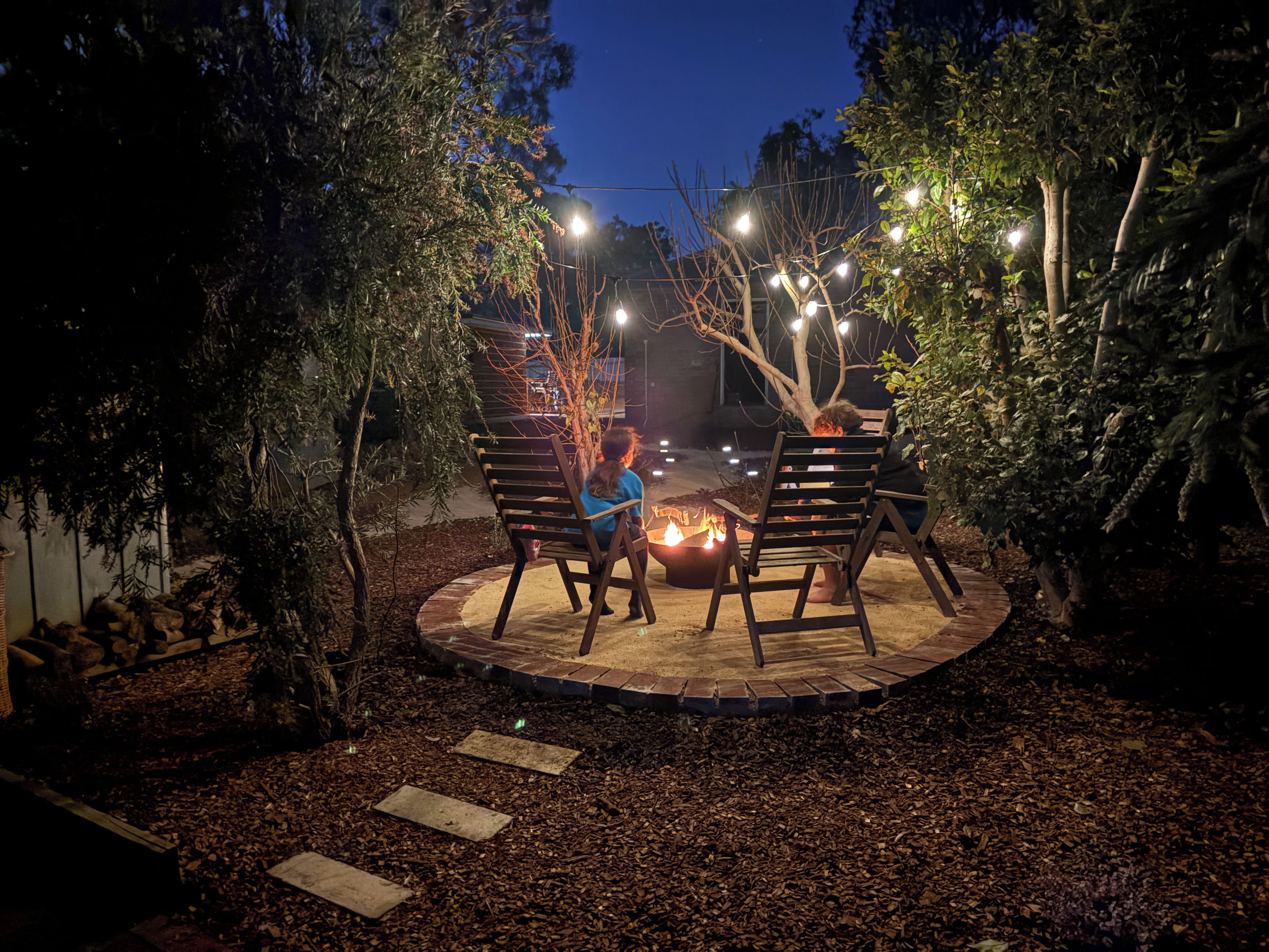 Un área de patio al aire libre con un brasero, sillas de madera y luces colgantes.
