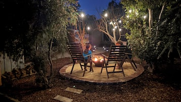 Un área de patio al aire libre con un brasero, sillas de madera y luces colgantes.