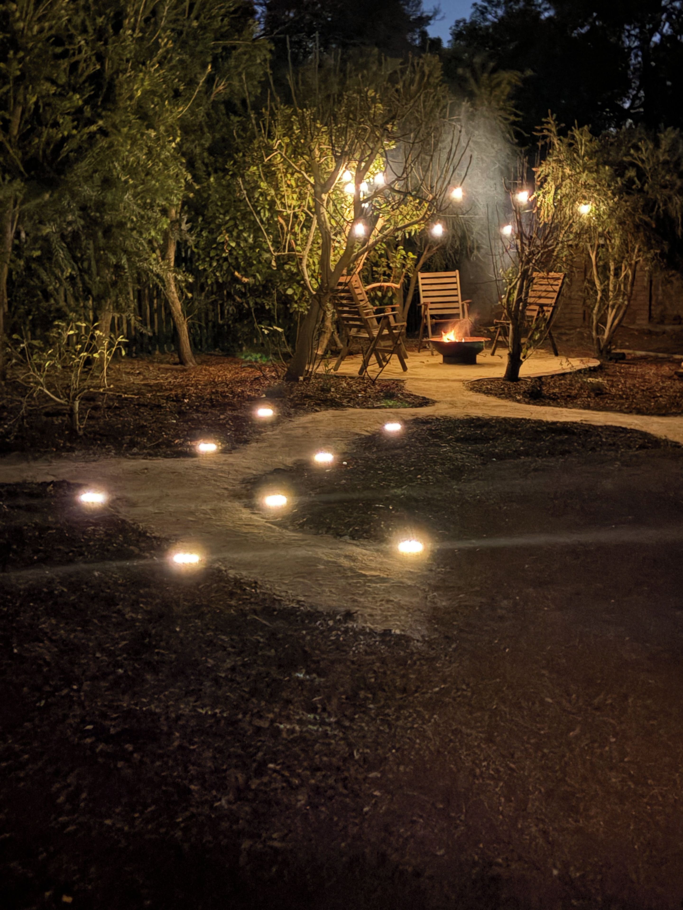 A pathway leading to a seating area with chairs and a fire pit, surrounded by trees and illuminated by string lights.