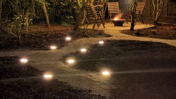 A pathway leading to a seating area with chairs and a fire pit, surrounded by trees and illuminated by string lights.