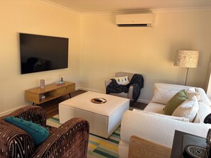 A living room with a flat-screen TV, a wooden side table, a white sofa, a wicker chair, and a floor lamp.