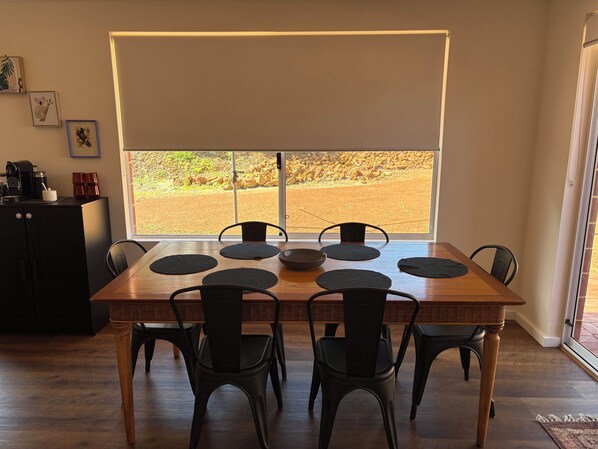 A dining area with a wooden table and chairs, a large window with a view of a rocky landscape, and a kitchenette with a coffee machine. - Grove Road Retreat - walk to Lesmurdie Falls! (Lesmurdie)