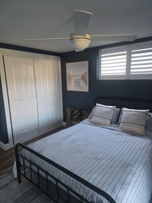 Ein Schlafzimmer mit einem Bett, einem Deckenventilator, einem Fenster mit Jalousien und einer Tür.