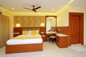 A hotel room with a bed, a desk, a chair, a wardrobe, and a ceiling fan. - Capital O 14702 Kovilakam Residency (Thrissur)