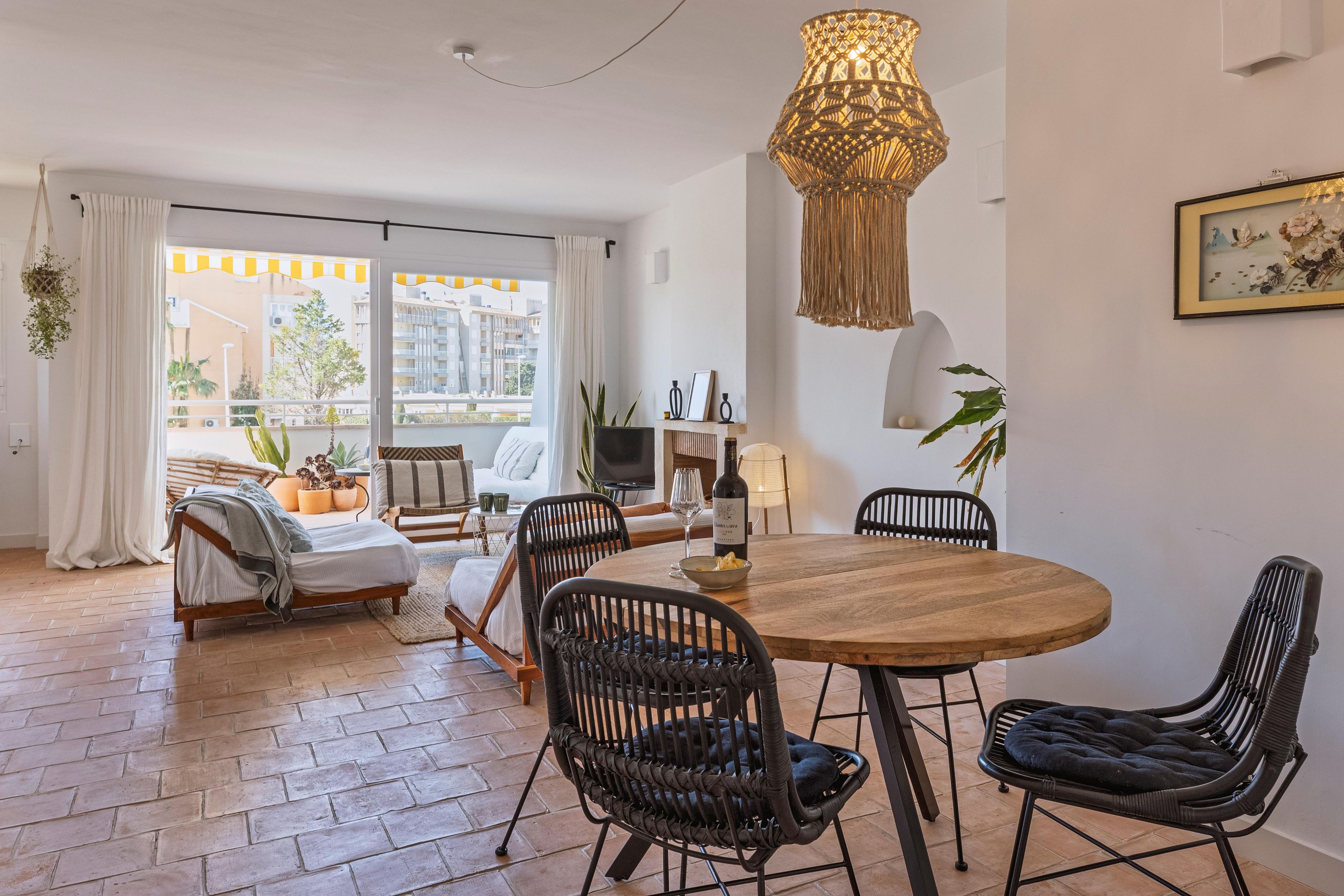 Ein modernes Wohnzimmer mit einem Holzesstisch, schwarzen Korbstühlen, einem großen Fenster mit Vorhängen und einer Hängelampe.