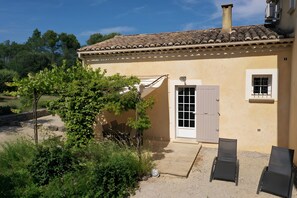 Un bâtiment avec un toit recouvert de tuiles, une porte et une fenêtre. Il y a un jardin avec de la verdure et un banc.