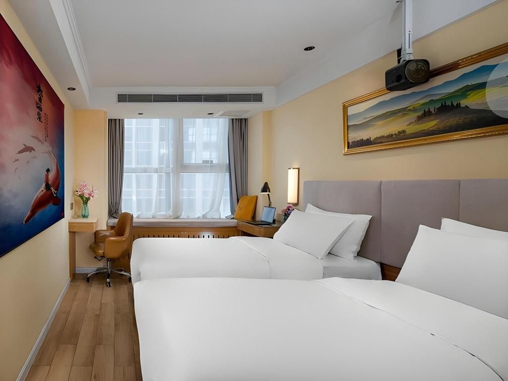 A hotel room with a large bed, a desk, a chair, a window with curtains, a painting on the wall, and a projector mounted on the ceiling.