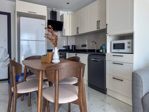 A modern kitchen with white cabinets, a Bosch dishwasher, a microwave, and a dining area with wooden chairs and a vase. - Qoople Elize B20 - Cozy 1-bedroom apartment in Alanya (Alanya)
