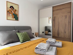 A hotel room with a bed, pillows, a green throw pillow, a yellow blanket, and two towels with a logo. - Qoople Elize B20 - Cozy 1-bedroom apartment in Alanya (Alanya)