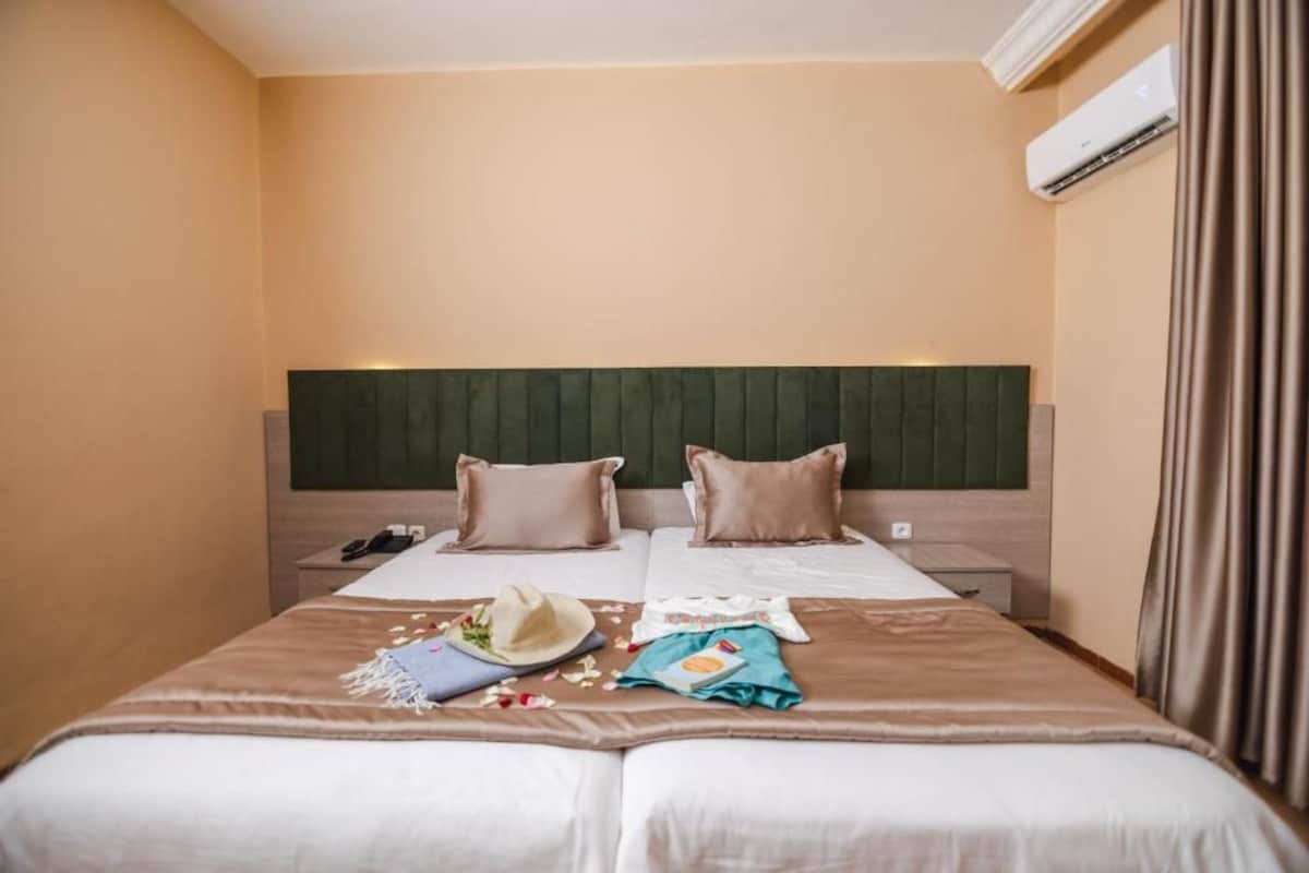 A hotel room with a bed, bedside tables, an air conditioning unit, and a curtain.