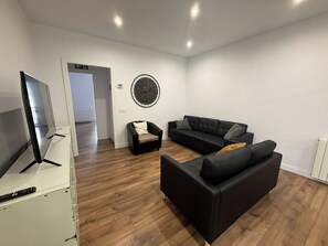 Ein modernes Wohnzimmer mit einem Flachbildfernseher, einer schwarzen Couch, einem kleineren Sessel und einer runden Wanduhr.