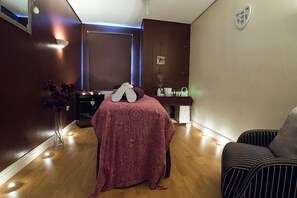 Una habitación con una cama cubierta por una manta estampada, una mesita de noche con una lámpara, una silla y un calentador fijado a la pared.