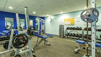 Un gimnasio con equipamiento de levantamiento de pesas, bancos y un estante de mancuernas en la pared.