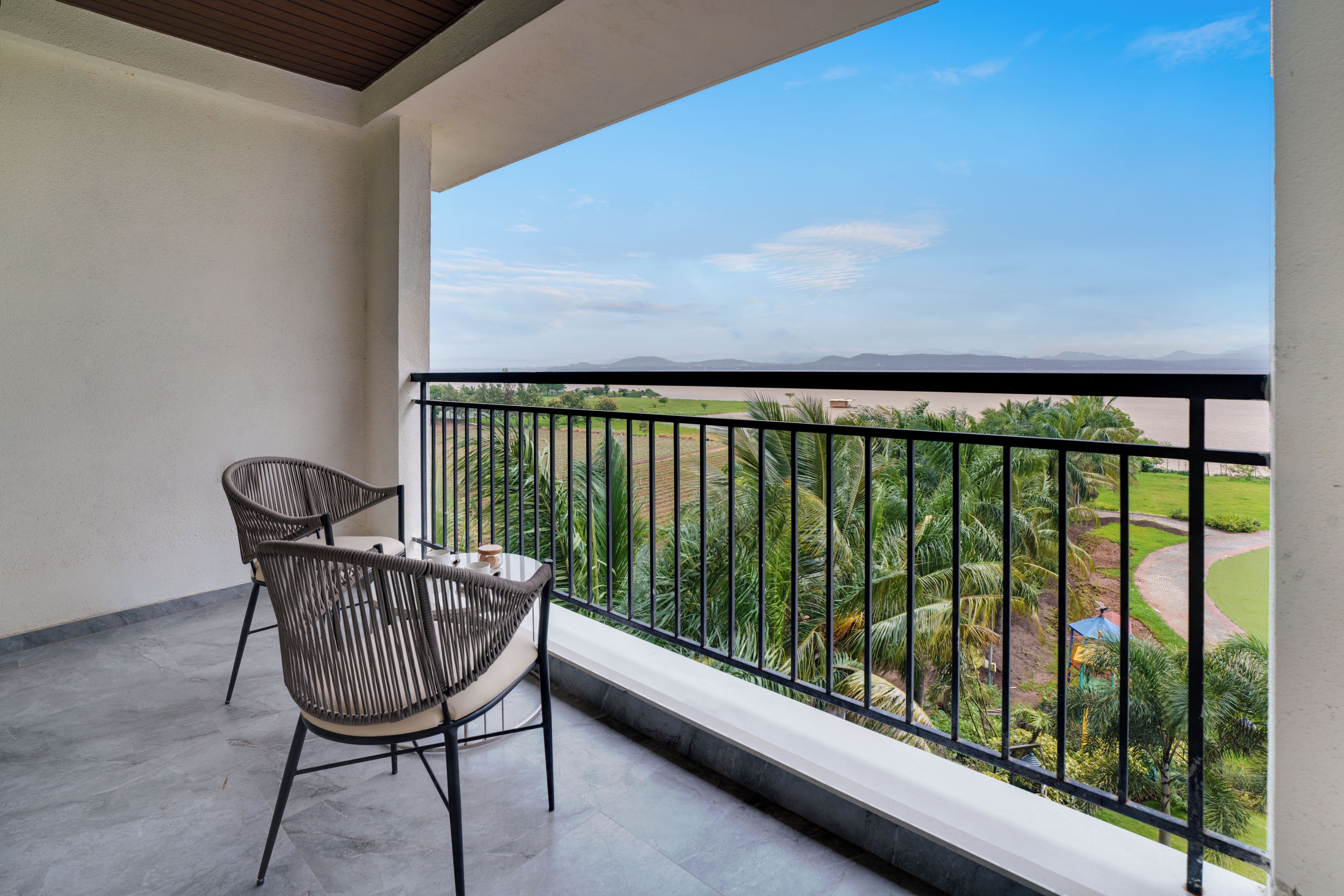 A balcony with two chairs and a table, overlooking a landscaped area with palm trees and a view of distant hills.