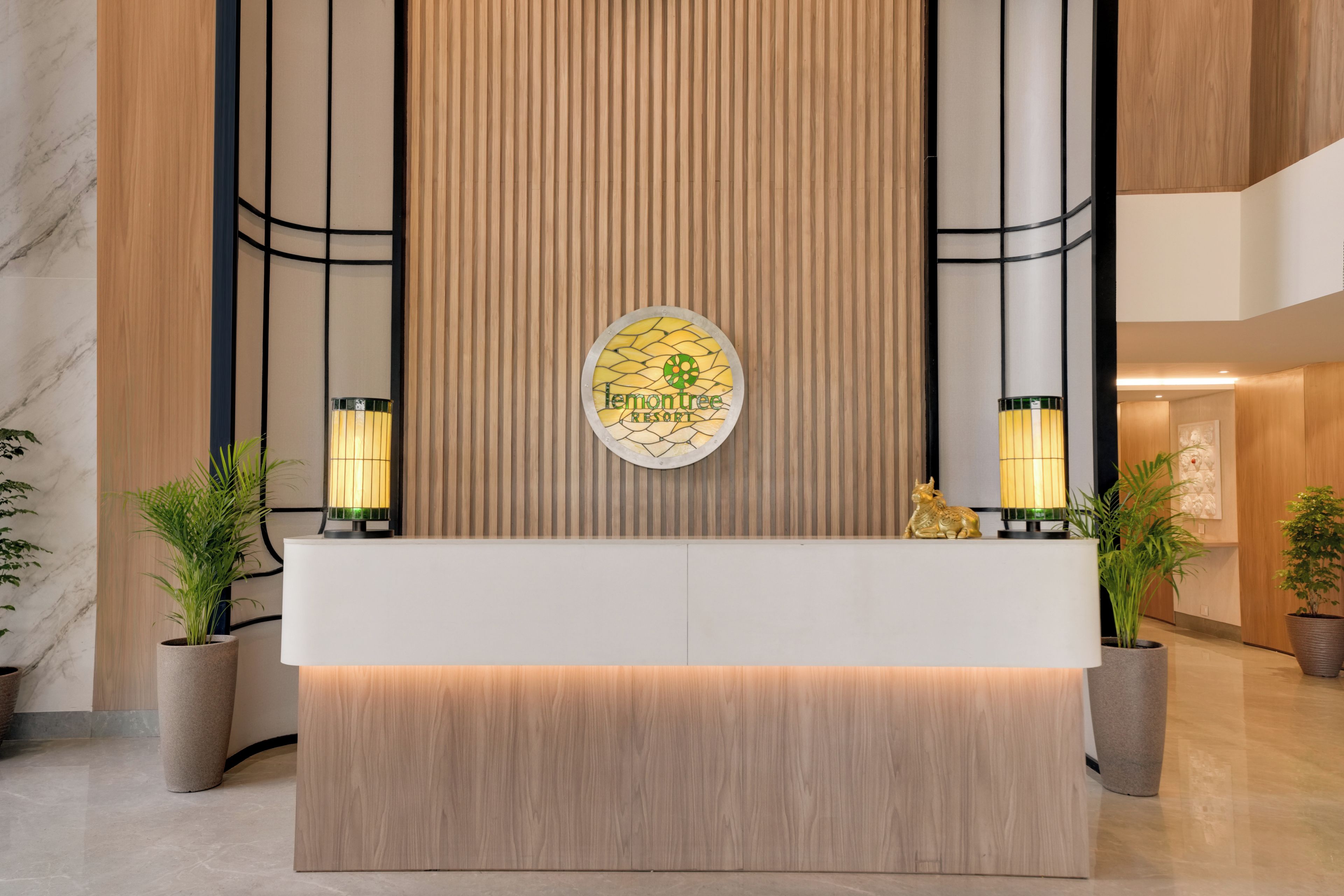 Un mostrador de recepción con un logotipo, dos lámparas de pared y plantas en macetas.