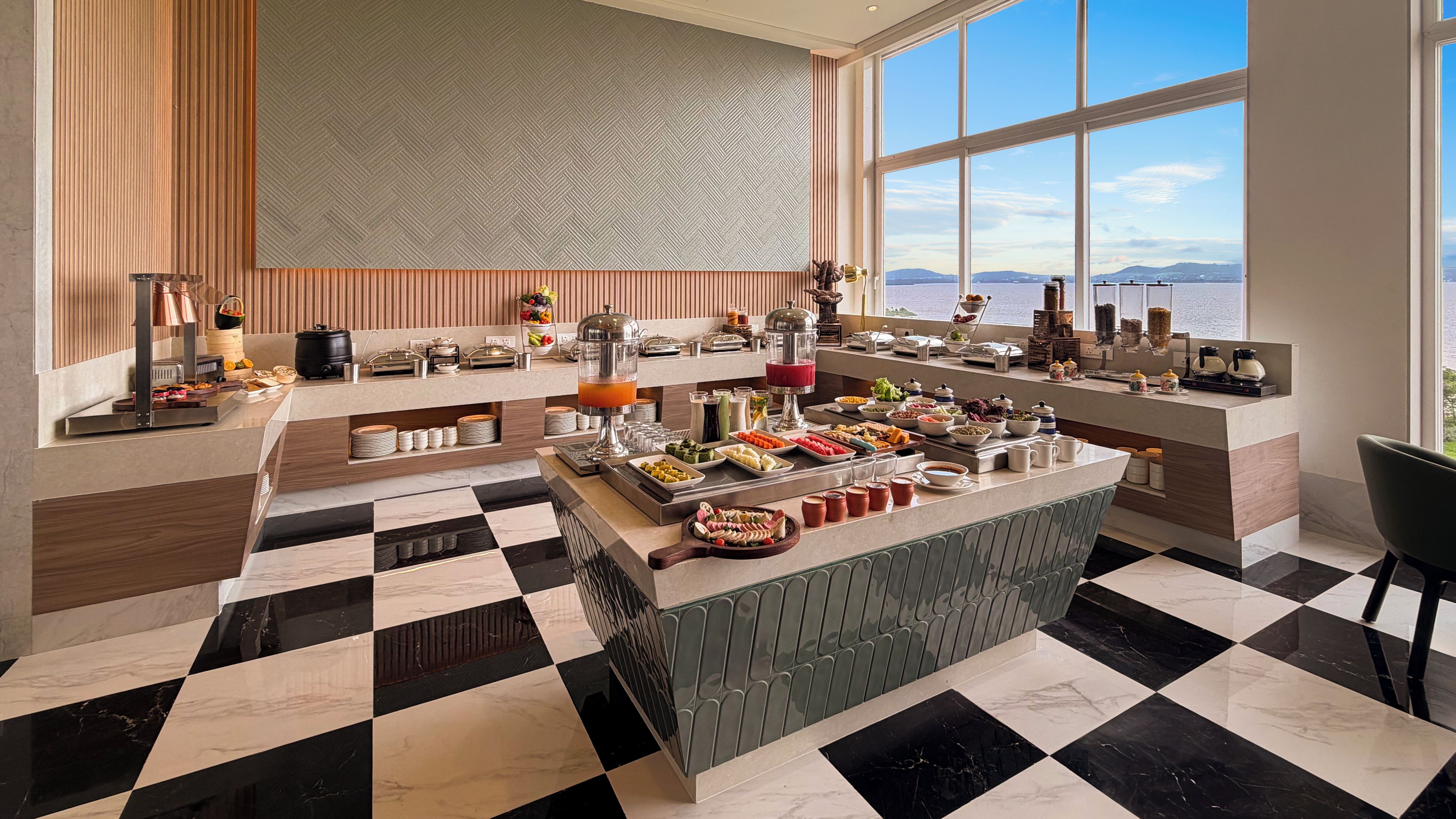 Un bufé de desayuno moderno con diversas opciones de comida, piso a cuadros y un amplio ventanal con vista al mar.