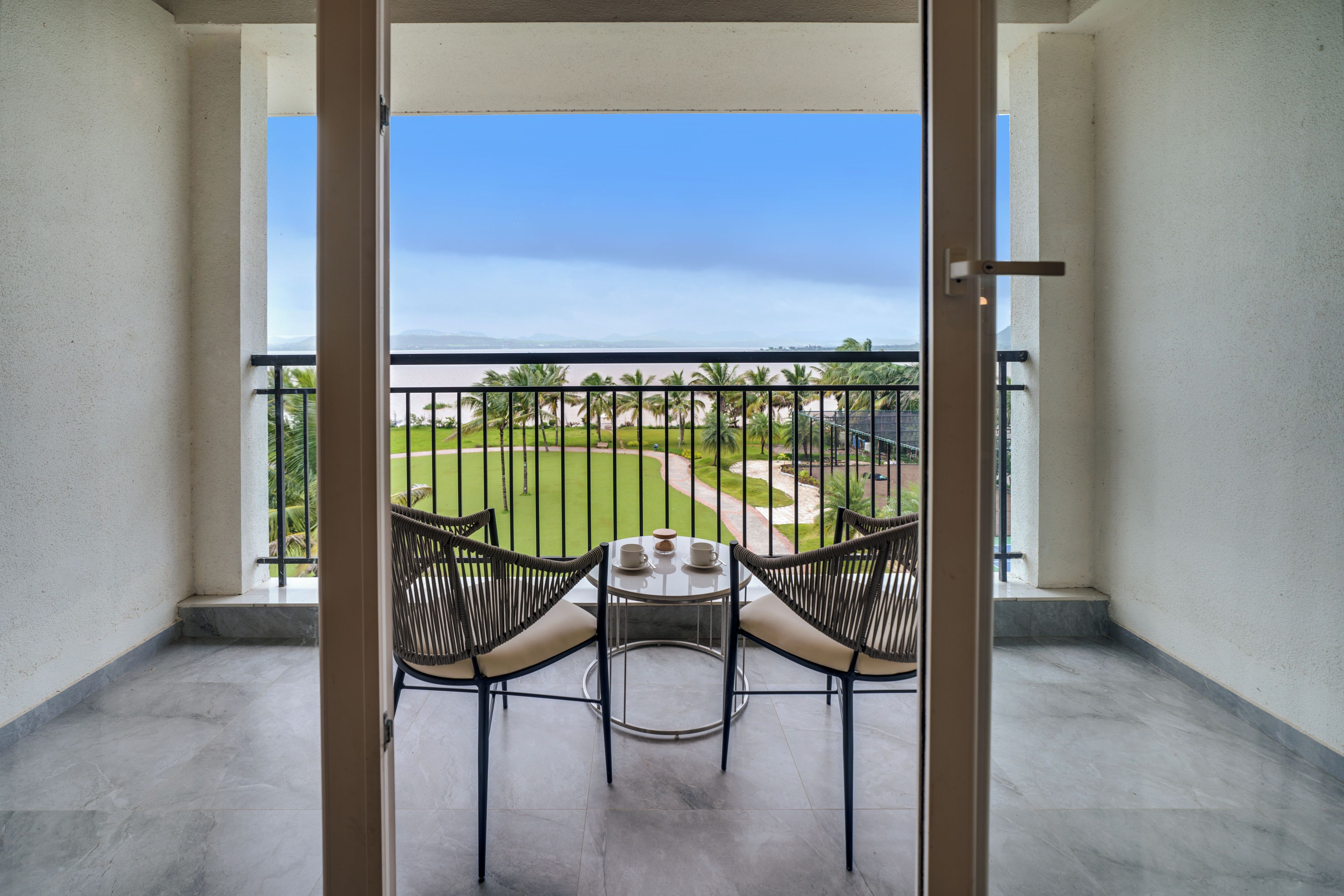 A balcony with two wicker chairs and a small table, overlooking a well-maintained lawn and a distant view of a body of water.
