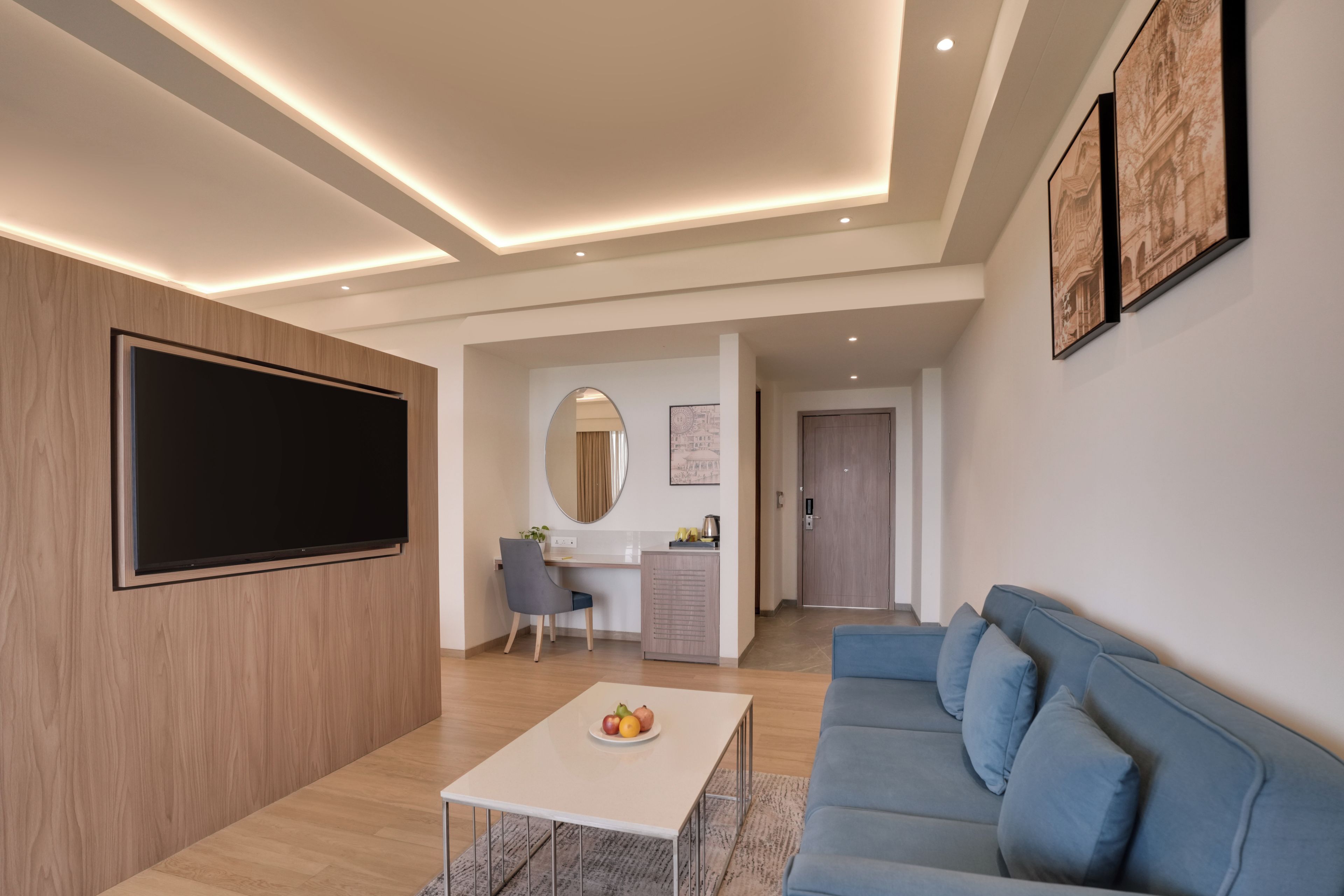 Een moderne woonkamer met een flatscreen televisie, een blauwe bank, een bijzettafeltje met fruit en een bureau met een spiegel en plant.