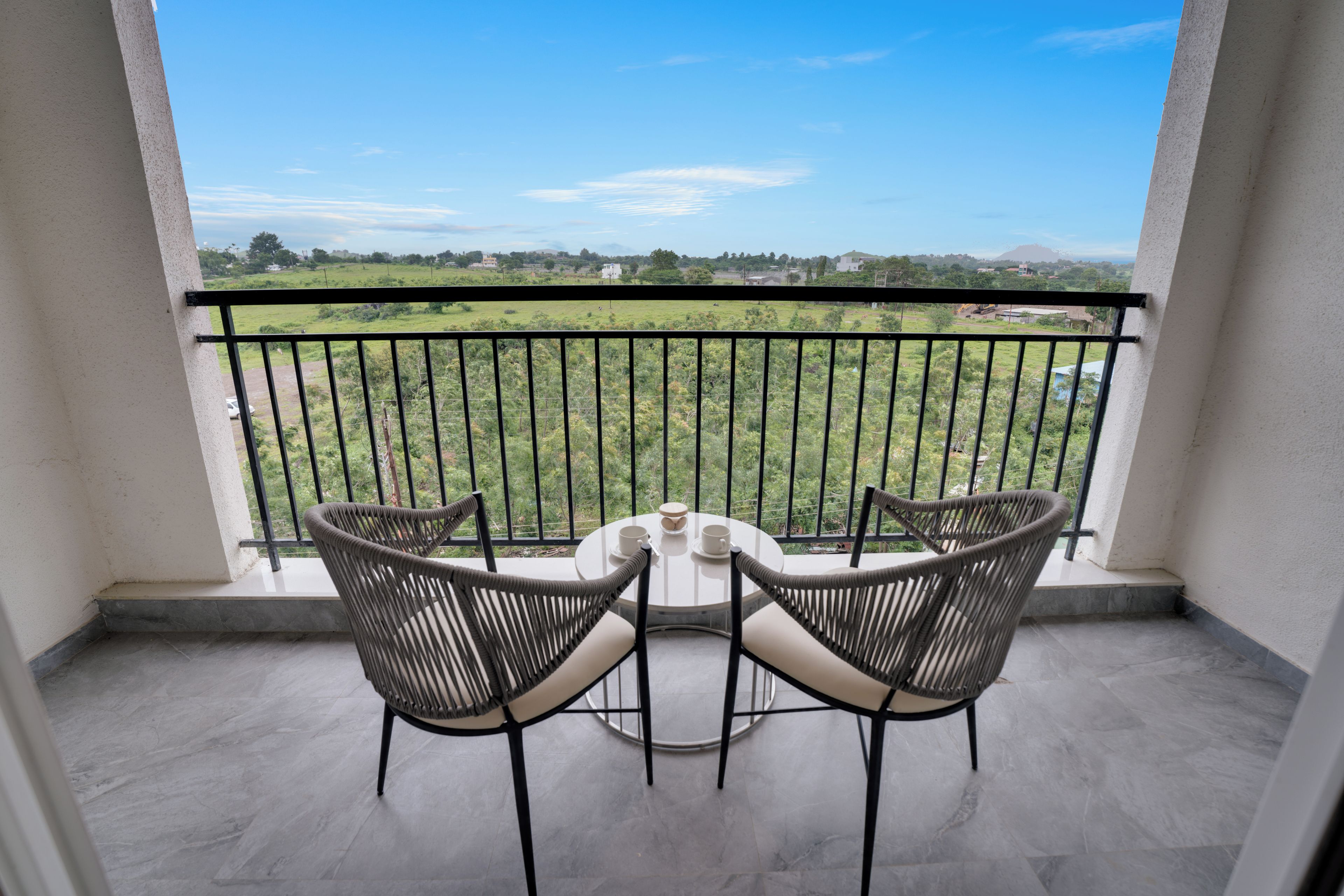 A balcony with two wicker chairs and a small table, overlooking a green landscape with a clear sky.