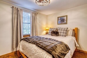 Un dormitorio con cama, cortinas, una lámpara, un cuadro enmarcado y suelo de madera.