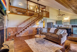 A spacious living room with a wooden staircase, a cozy fireplace, and a kitchen area. - Canopy View Lodge - Awesome Mountain Views, Hot Tub!!! (Sevierville)