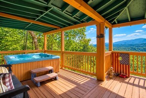 Una terraza de madera con jacuzzi, asientos y vistas a una cadena montañosa boscosa.