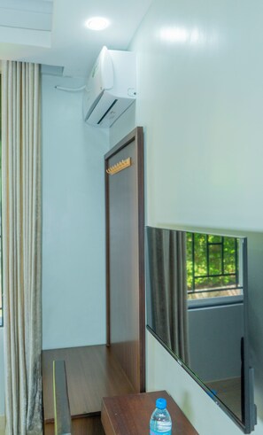A hotel room with a wooden door, a small table with a water bottle, and a window with a view of greenery.