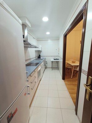 A narrow kitchen with a refrigerator, stove, and sink. There is a small table with chairs in the adjacent dining area. - Alojamiento Pl. del Ángel junto Pl. Mayor (Salamanca)