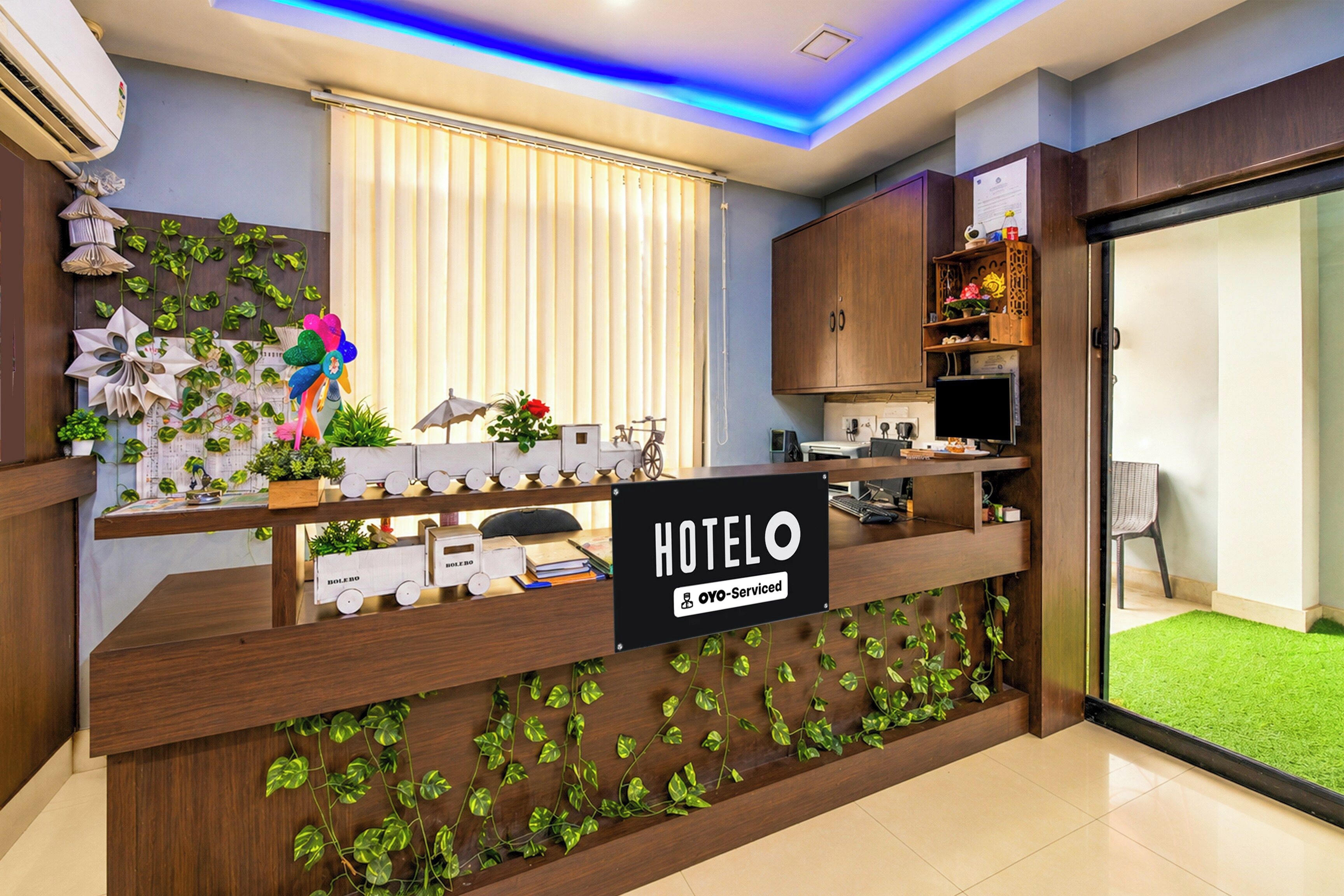 Ein Hotelrezeptionstresen, erkennbar als von OYO bewirtschaftet. Ein Fenster mit Jalousien, eine Pflanzenwand und ein kleiner Schreibtisch mit Computer und Stuhl sind vorhanden.