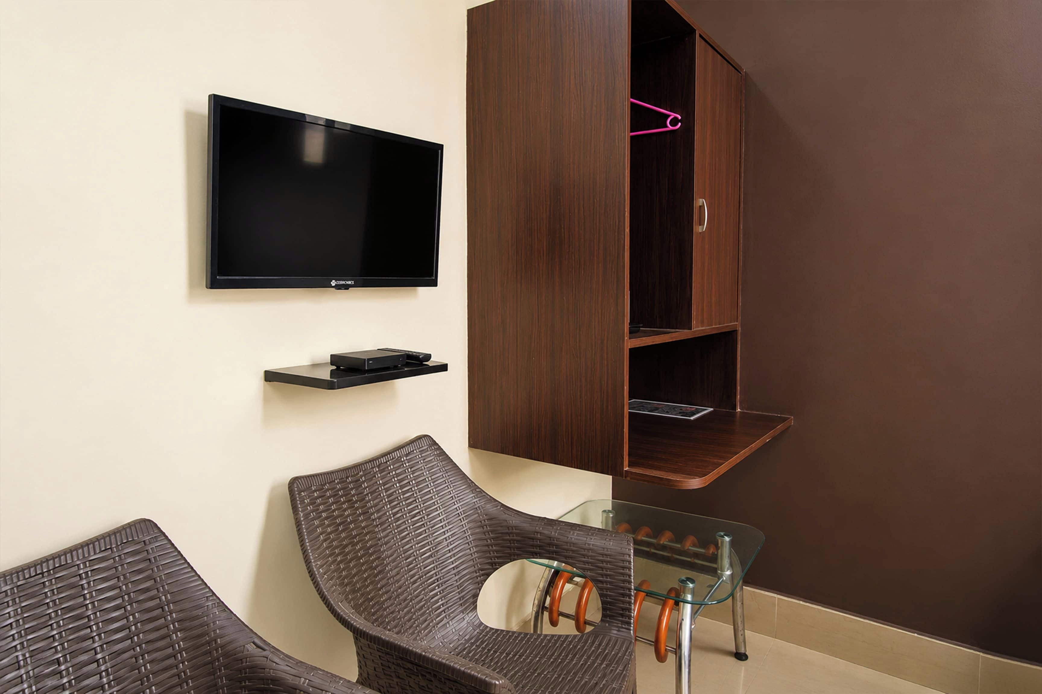 Una habitación con una televisión de pantalla plana montada en la pared, un armario de madera con un perchero rosa y dos sillas de mimbre con una mesa de vidrio.