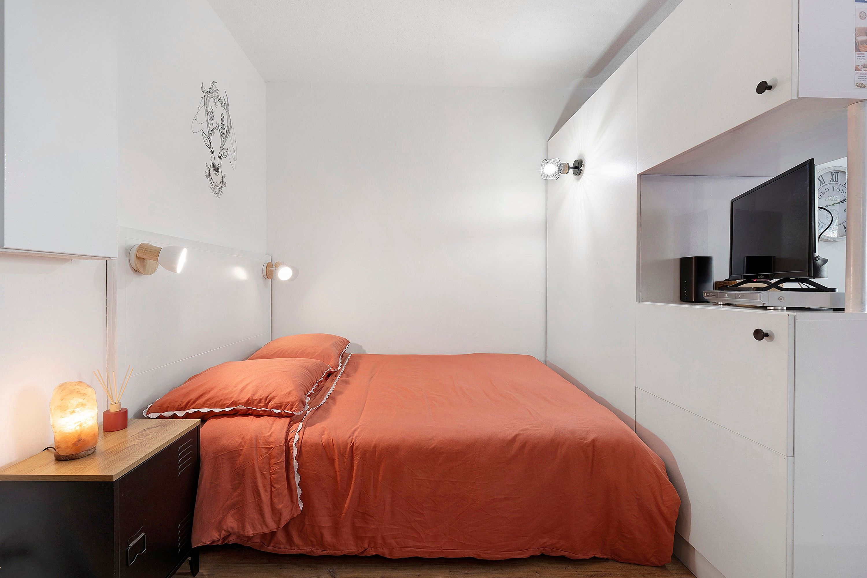 Un dormitorio con cama, mesita de noche, lámpara y televisor sobre un mueble.