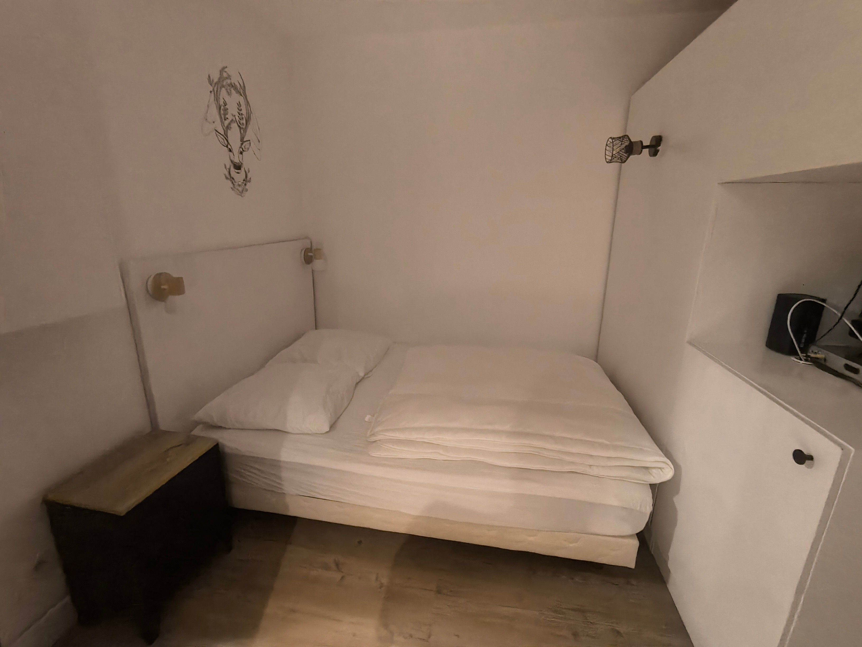 A small, single-bed room with a wooden nightstand, a wall-mounted shelf, and a bed with white bedding.